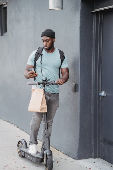 A young man with medium-dark skin, wearing a dark beanie, light blue t-shirt, and grey jeans, stands outdoors against a grey concrete wall, holding a smartphone in his right hand. He carries a black b
