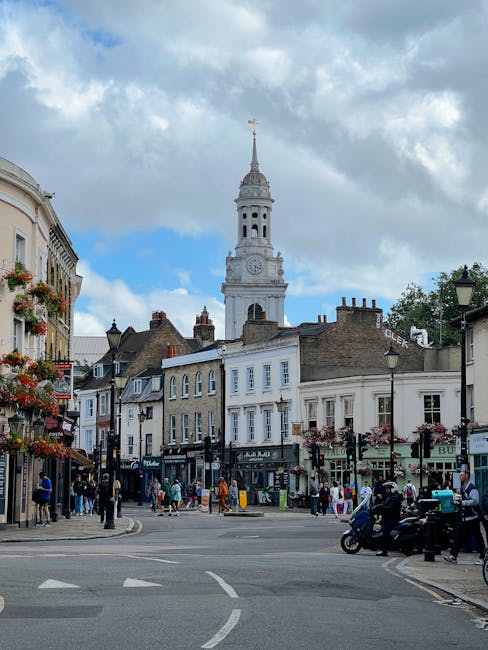 A vibrant street scene near Cheshunt High Street featuring a historic white clock tower with a golden weathervane on top, surrounded by old-fashioned buildings with decorative shopfronts and hanging f