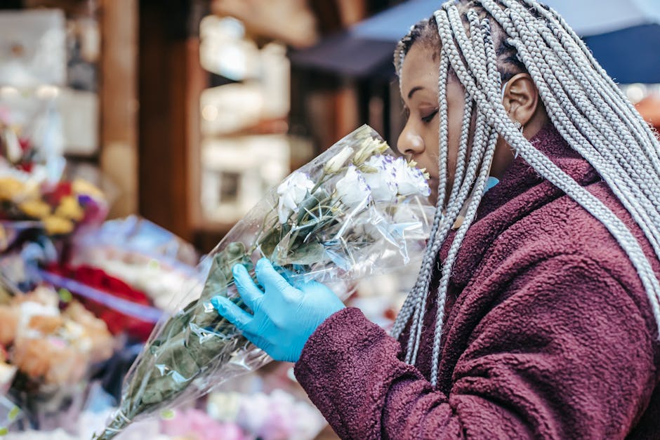 A woman with intricately styled silver braids is holding a large bouquet of fresh white roses wrapped in clear cellophane, examining the flowers closely. She is dressed in a dark burgundy fleece jacke